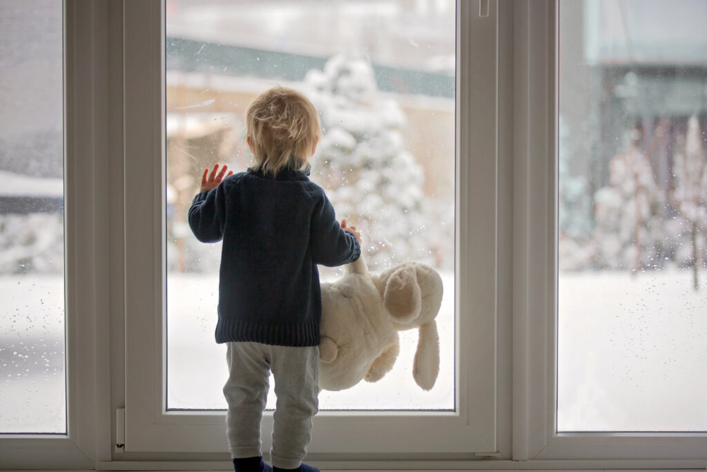 Toddler child standing in front of a big french doors, leaning against it looking outside at a snowy nature, holding bunny toy